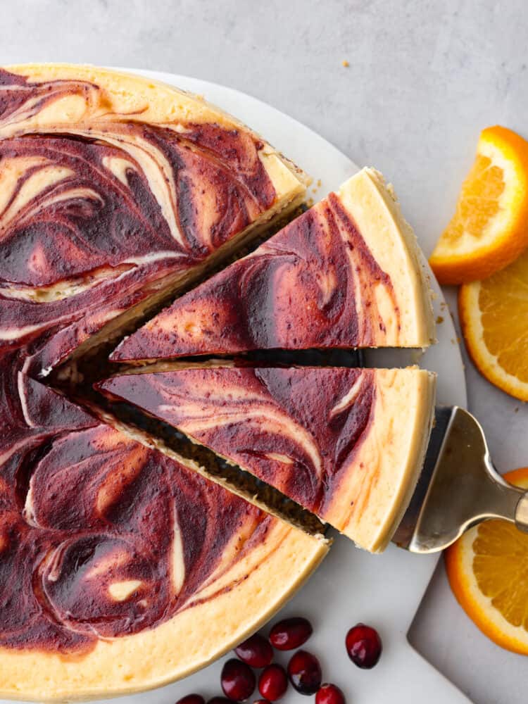 There is no better way to ring in the holidays than this Cranberry Orange Cheesecake. It's infused with a cranberry sauce that takes this dessert to another level, and is sure to be the talk of your party! A whole orange cranberry cheesecake with two slices slightly removed on a plate with oranges and cranberries to the side.