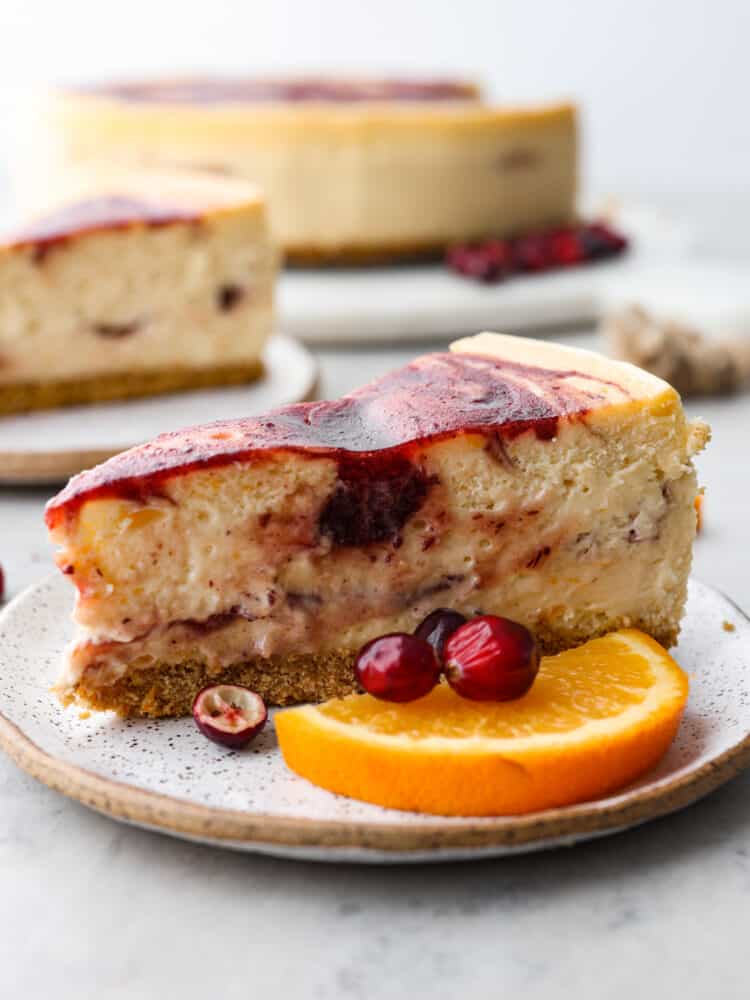 There is no better way to ring in the holidays than this Cranberry Orange Cheesecake. It's infused with a cranberry sauce that takes this dessert to another level, and is sure to be the talk of your party! Cheesecake on a plate with cranberries and oranges in front of it.