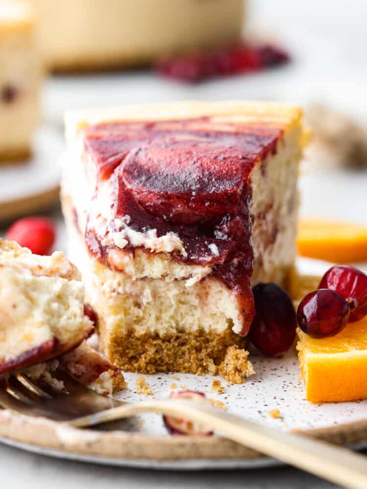 There is no better way to ring in the holidays than this Cranberry Orange Cheesecake. It's infused with a cranberry sauce that takes this dessert to another level, and is sure to be the talk of your party! A slice of cheesecake with a bite taken out of it with a fork and fresh fruit next to it.