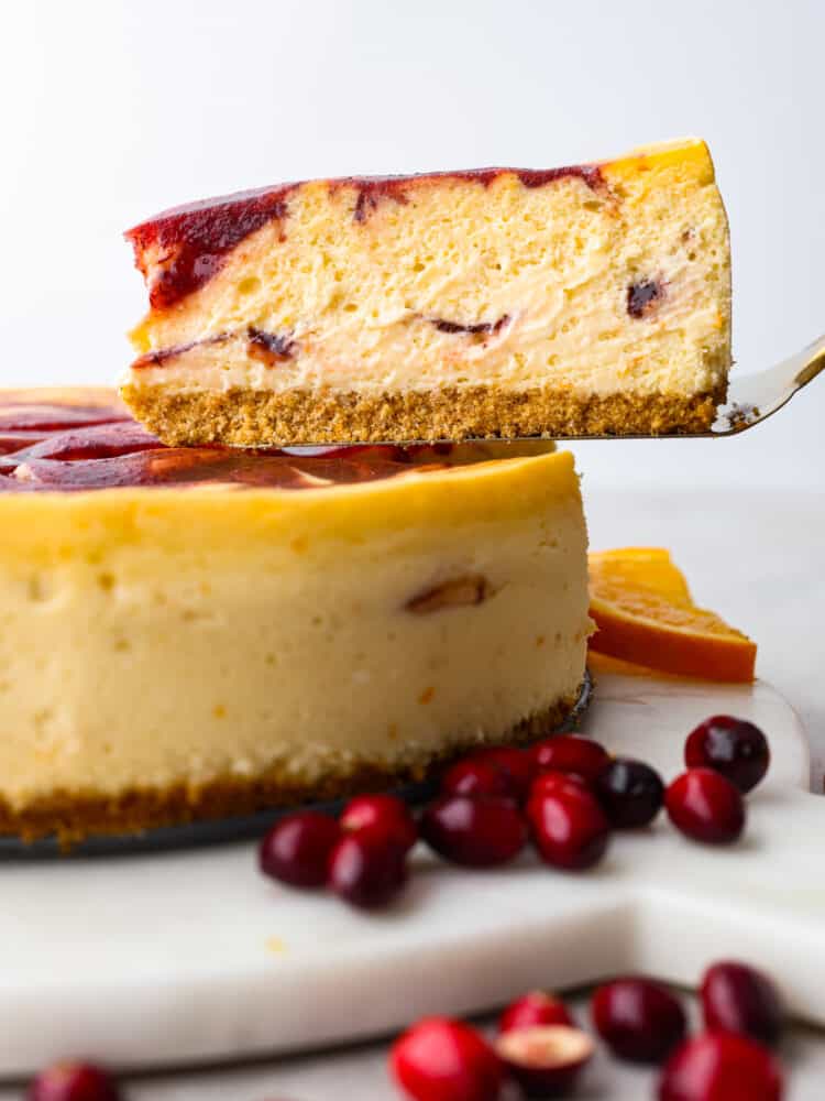 There is no better way to ring in the holidays than this Cranberry Orange Cheesecake. It's infused with a cranberry sauce that takes this dessert to another level, and is sure to be the talk of your party! A piece of orange cranberry cheesecake being lifted out of the whole by a pie spatula.