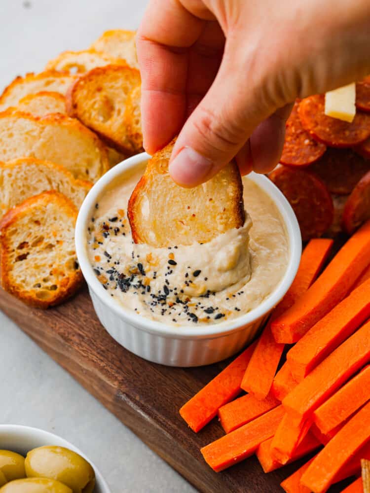 This football charcuterie board is perfect for any of your game day or tailgate parties! It's packed with all of the perfect finger food that everyone will love and you will be making this over and over during football season! A slice of crostini being dipped in some hummus on the charcuterie board.