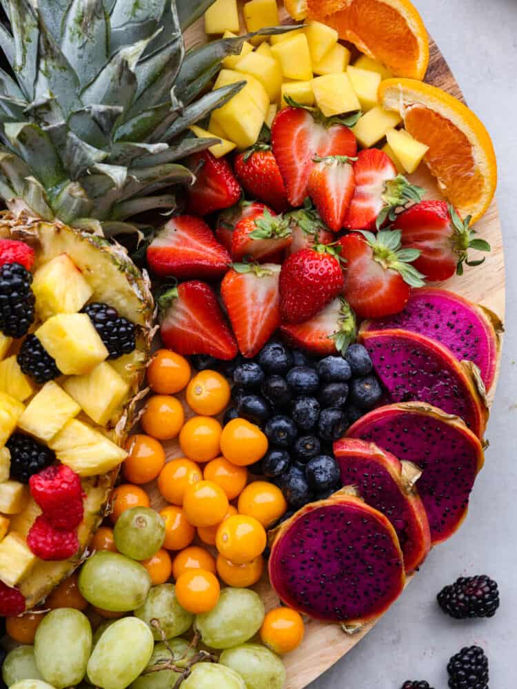 You will love the vibrant colors of all your favorite fruits beautifully displayed together on a fruit charcuterie board. Serve this as a perfect side dish or dessert at your next party alongside a creamy fruit dip! Close overhead view of the grapes, kumquat, dragonfruit, berries, pineapple, and orange slices on the charcuterie board.