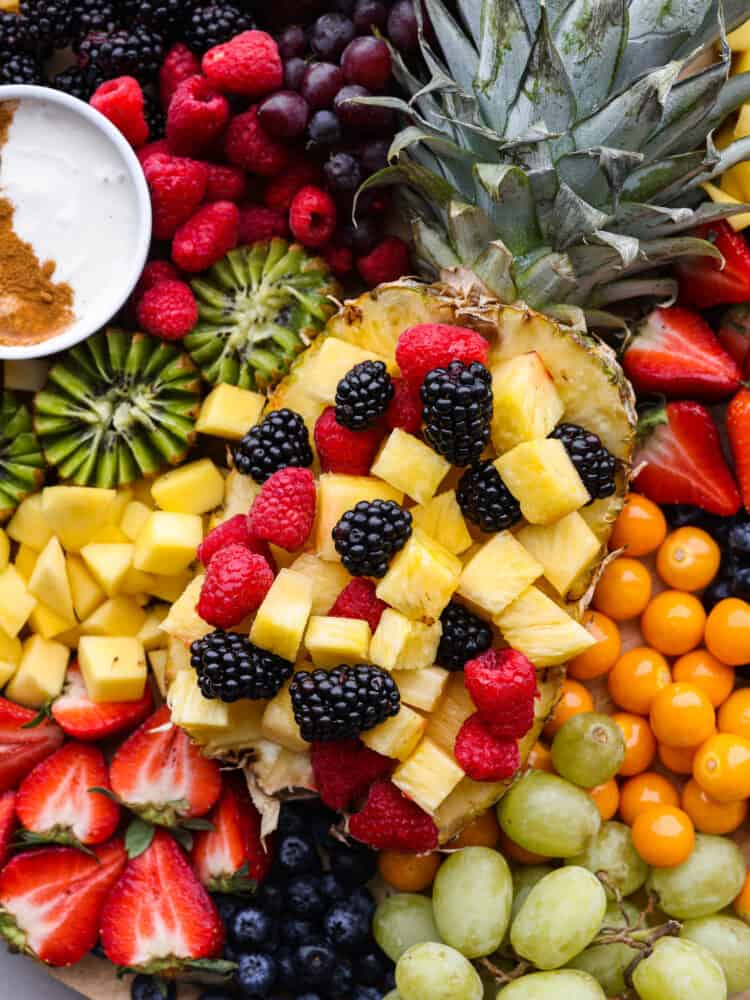 You will love the vibrant colors of all your favorite fruits beautifully displayed together on a fruit charcuterie board. Serve this as a perfect side dish or dessert at your next party alongside a creamy fruit dip! Close overhead view of the sliced whole pineapple filled with pineapple chunks and berries. The rest of the fruit is scattered around the pineapple with fruit dip on the side.