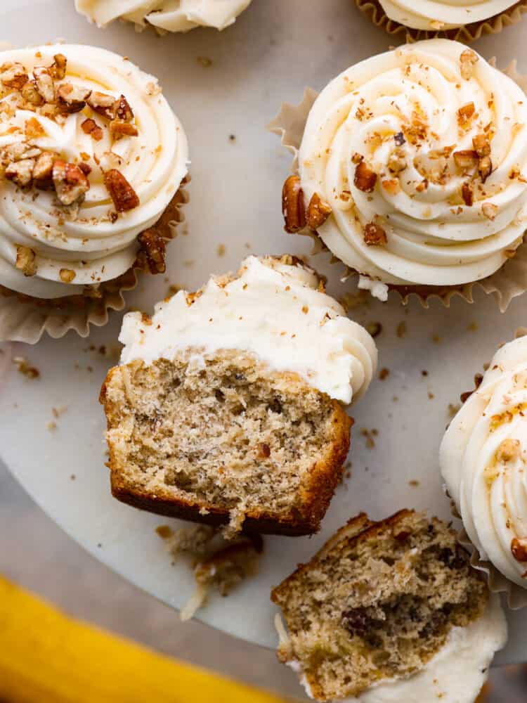 A bite-sized take on a classic Southern dessert, these hummingbird cupcakes are sure to win your heart! Filled with pecans, chopped fruit, and topped with smooth cream cheese frosting, you won't be able to stop at one! Top-down view of hummingbird cupcakes on a gray serving tray. One is cut in half.