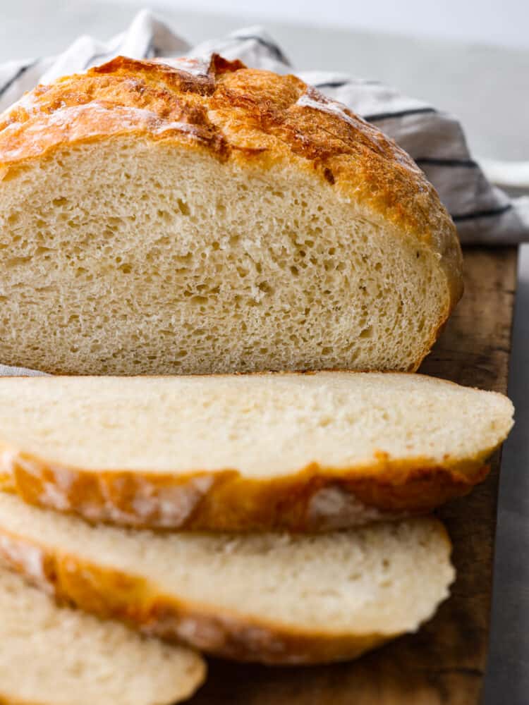Potato bread is a soft white bread that is spongy and delicious. The potatoes add a fluffy yet firm texture making it perfect for sandwiches, toast, or warmed with jam. A cut loaf of potato bread on a rustic cutting board. A striped kitchen towel is next the the loaf of bread.