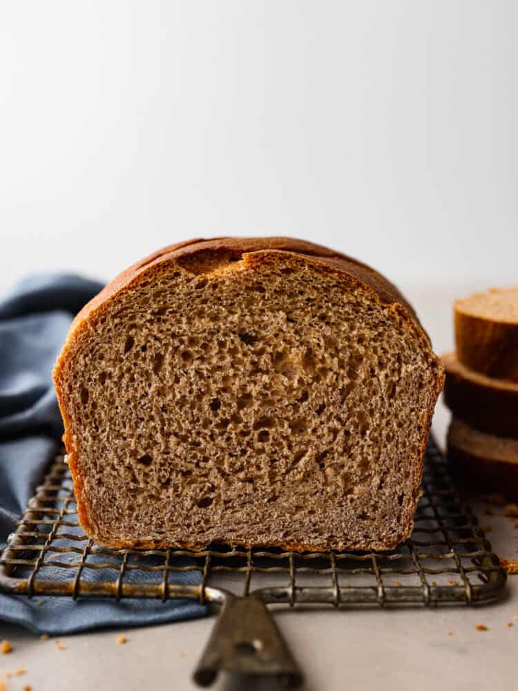 This Sprouted Wheat Bread is fluffy, moist, and full of rich flavor! If you are a skeptic of whole wheat bread because you've tried store-bought in the past, get ready to change your mind! I promise even picky eaters love the nutty flavor and soft texture of this homemade bread. Whole wheat bread loaf sliced open facing the viewer sitting on a wire rack with stacked bread in the background.