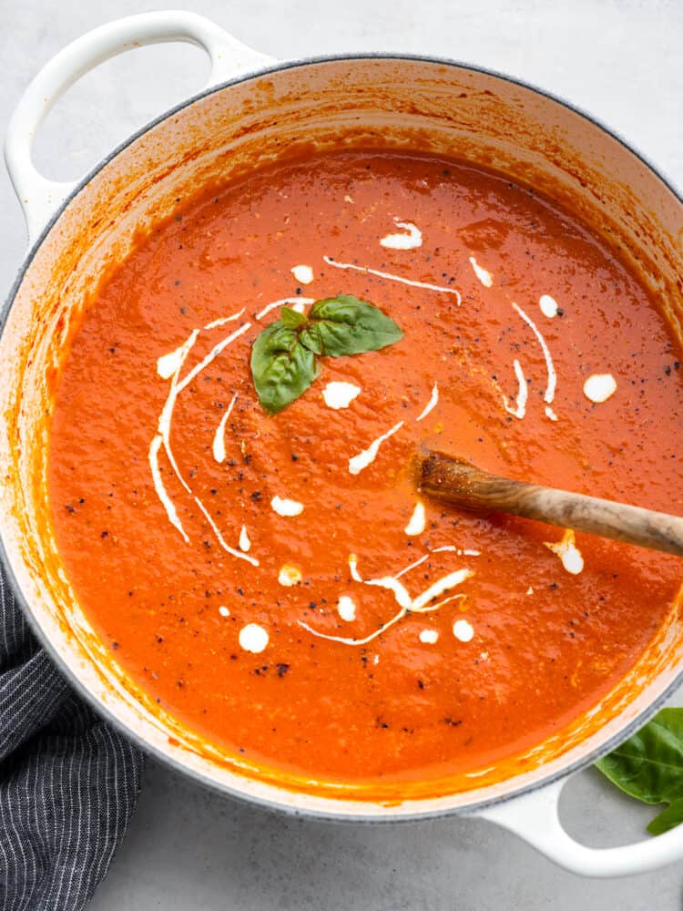 Tomato basil soup is a healthy classic that's simple to make from scratch and full of flavor! It only takes just over half an hour. The whole family will enjoy it as the temperatures drop and the evenings become crisper. Top-down view of tomato basil soup in a blue pot, garnished with herbs and a dollop of cream.