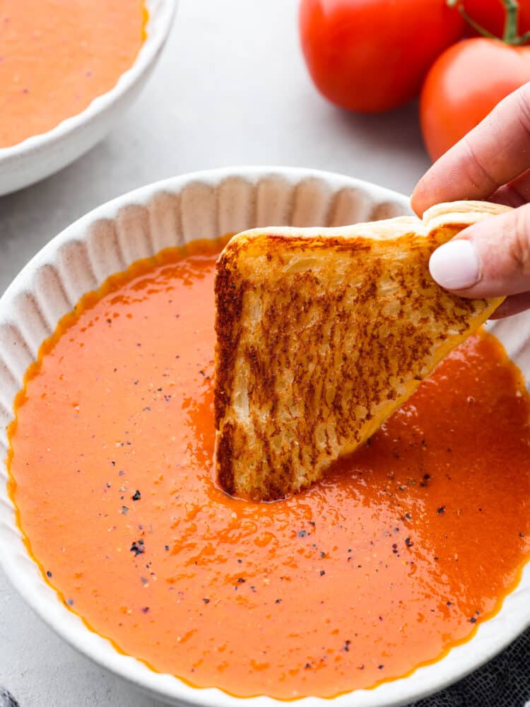 Homemade Tomato Soup is creamy, flavorful, and the ultimate fall dish. Ripe tomatoes flavored with garlic and onion are blended with chicken broth and cream to make the most flavorful homemade tomato soup you'll ever try! Tomato soup in a white bowl with a hand dipping a piece of toast into it.