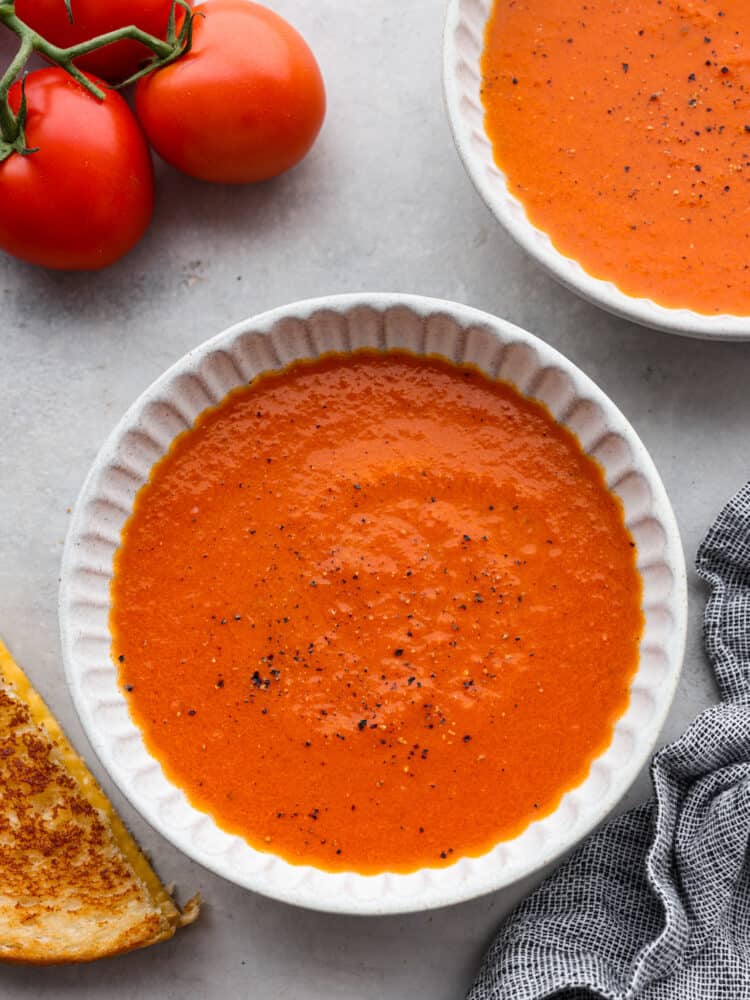 Homemade Tomato Soup is creamy, flavorful, and the ultimate fall dish. Ripe tomatoes flavored with garlic and onion are blended with chicken broth and cream to make the most flavorful homemade tomato soup you'll ever try! Two bowls of tomato soup with tomatoes off to the side.