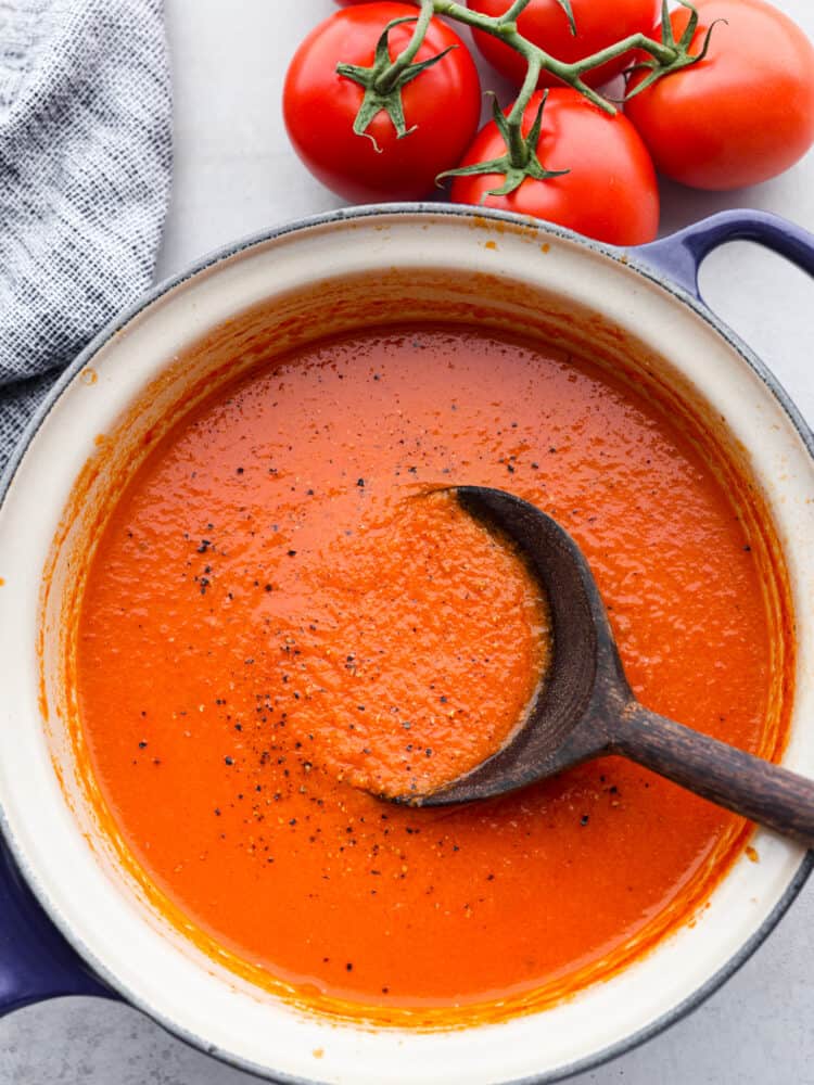 Homemade Tomato Soup is creamy, flavorful, and the ultimate fall dish. Ripe tomatoes flavored with garlic and onion are blended with chicken broth and cream to make the most flavorful homemade tomato soup you'll ever try! Tomato soup with a black wooden spoon sitting in it with ripe tomatoes and a blue tea towel to the side.