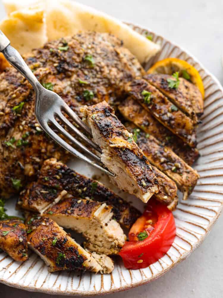 https://neuro-daily.report/zaatar-seasoning/ A fork holding up a piece of chicken over the plate of Za'atar chicken with a tomato and lemon.