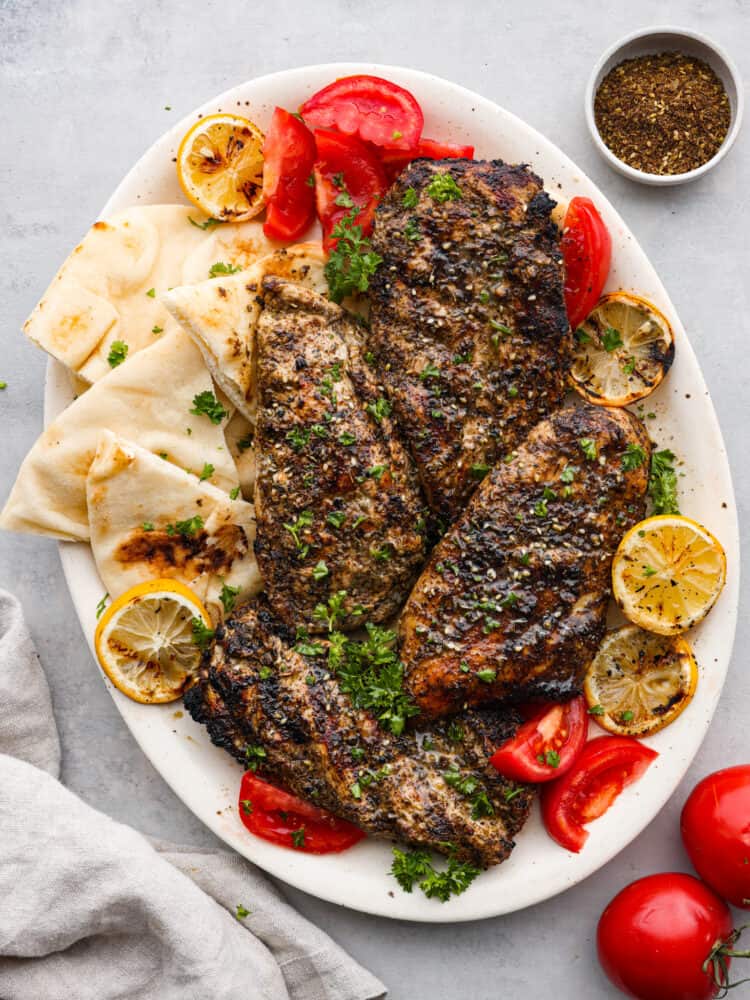 https://neuro-daily.report/zaatar-seasoning/ Za'atar chicken on a plate with tomatoes, lemons, pita bread, and seasoning to the side.