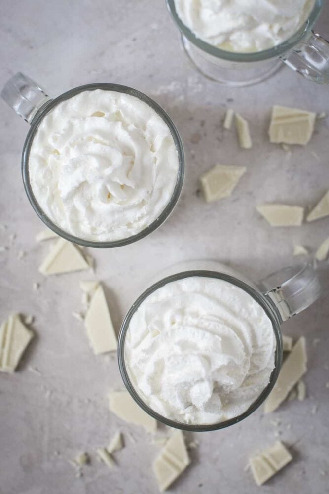 White Hot Chocolate is a rich and creamy decadent drink that melts in your mouth with a smooth, sweet white chocolate and vanilla flavoring. Warm up with this delicious hot chocolate throughout the entire holiday season! The top view of some white hot chocolate in glass cups with whipped cream on top.