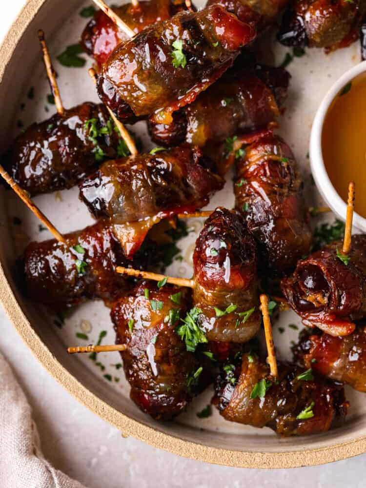 Bacon wrapped dates are the perfect combination of sweet and salty and will be the most talked about appetizer at your next party. Sweet tender dates wrapped in bacon and drizzled with honey and you won't believe how amazing these are! Close up view of bacon wrapped dates on a tan colored plate. A small bowl of honey is on the plate with a tan colored kitchen towel styled next to the plate. Chopped parsley is sprinkled on top.