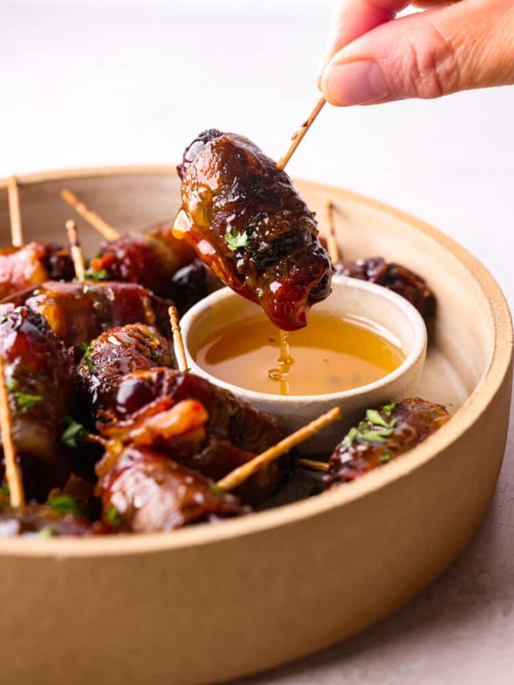 Bacon wrapped dates are the perfect combination of sweet and salty and will be the most talked about appetizer at your next party. Sweet tender dates wrapped in bacon and drizzled with honey and you won't believe how amazing these are! A close up side view of a bacon wrapped date being dipped into a small bowl of honey.