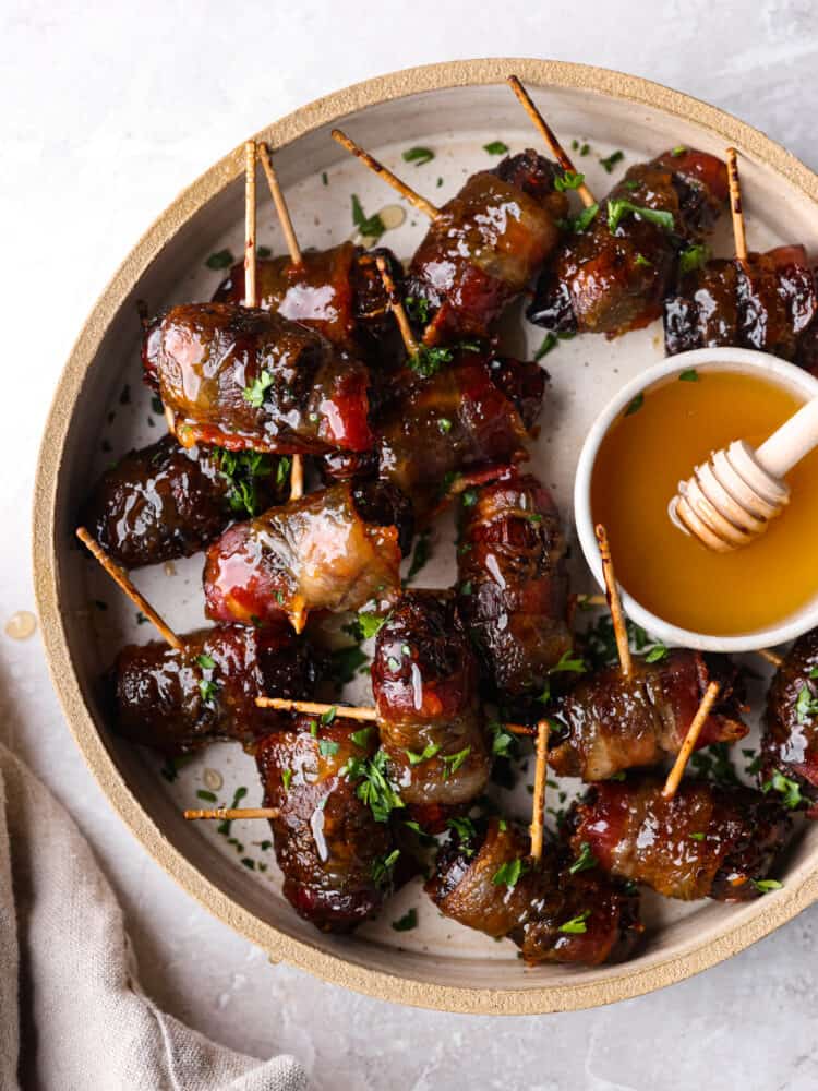 Bacon wrapped dates are the perfect combination of sweet and salty and will be the most talked about appetizer at your next party. Sweet tender dates wrapped in bacon and drizzled with honey and you won't believe how amazing these are! Top view of bacon wrapped dates on a tan colored plate. A small bowl of honey is on the plate with a tan colored kitchen towel styled next to the plate.