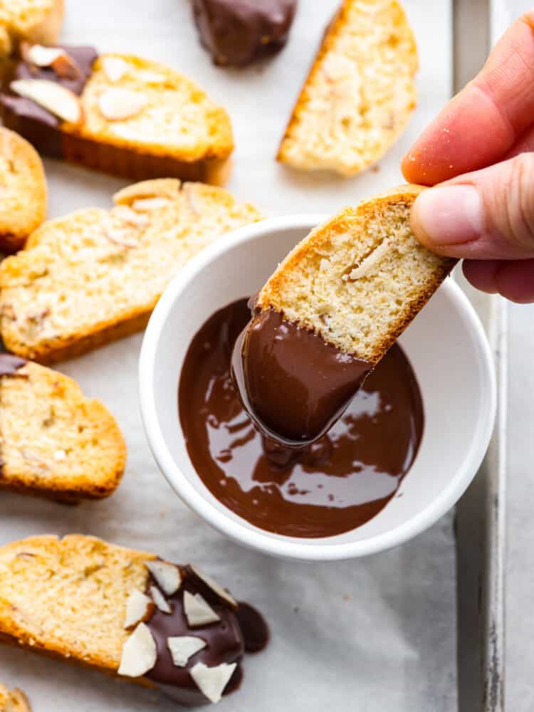 These Classic Biscotti are crispy and crunchy and melt in your mouth with a little help from some hot cocoa or coffee. Dipped in dark chocolate and sprinkled with sliced almonds, this fun treat is perfect for sharing with friends on a cold day! A hand dipping the biscotti in chocolate.