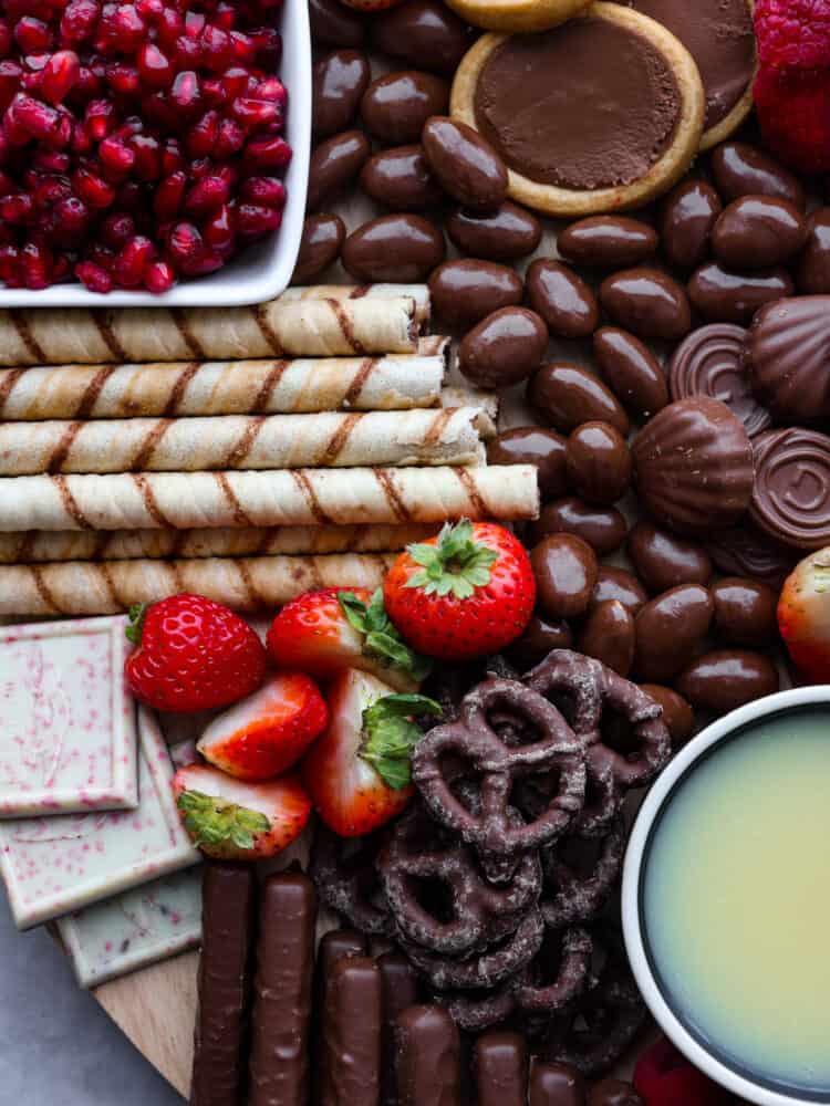 This chocolate charcuterie board is a chocolate lover's dream! Enjoy a chocolatey assortment of cookies, truffles, chocolate-covered pretzels, and fruit, along with decadent sauces for dipping! Close up photo of the chocolate charcuterie board. Cookies, chocolate almonds, and fruit are scattered on the board.