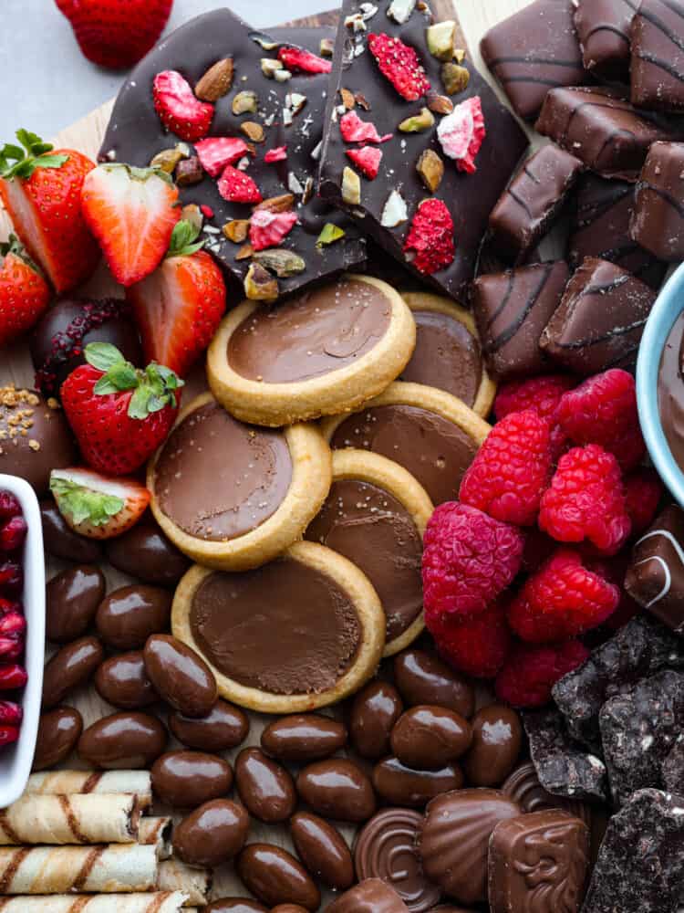 This chocolate charcuterie board is a chocolate lover's dream! Enjoy a chocolatey assortment of cookies, truffles, chocolate-covered pretzels, and fruit, along with decadent sauces for dipping! Close up photo of the chocolate charcuterie board. Cookies, chocolate almonds, and fruit are scattered on the board.