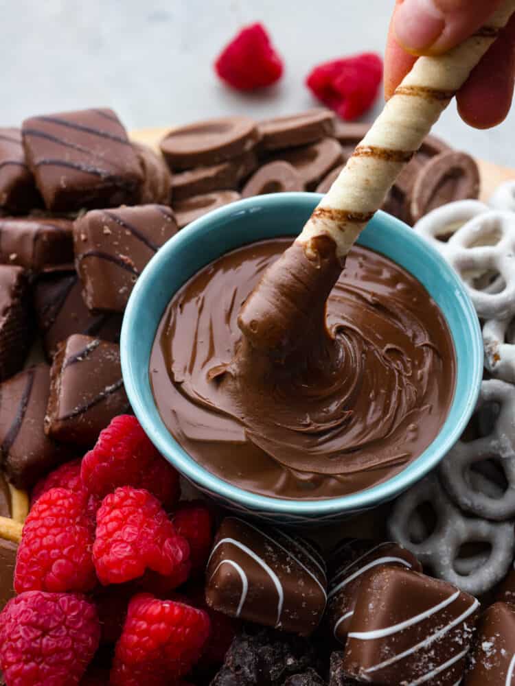 This chocolate charcuterie board is a chocolate lover's dream! Enjoy a chocolatey assortment of cookies, truffles, chocolate-covered pretzels, and fruit, along with decadent sauces for dipping! A close up photo of a cookie being dipped into a bowl of nutella. Chocolate, candy, and raspberries surround the bowl.