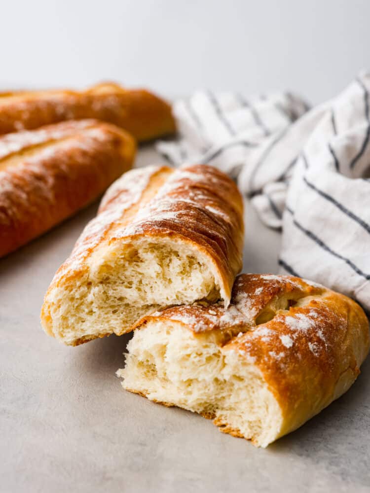Cuban bread is a delightfully soft and fluffy bread that is shaped like a French baguette. It has a soft crust and the secret ingredient is lard which is what gives this bread great flavor and the perfect chew! A loaf of bread ripped in half showing the inside with other loaves in the background.