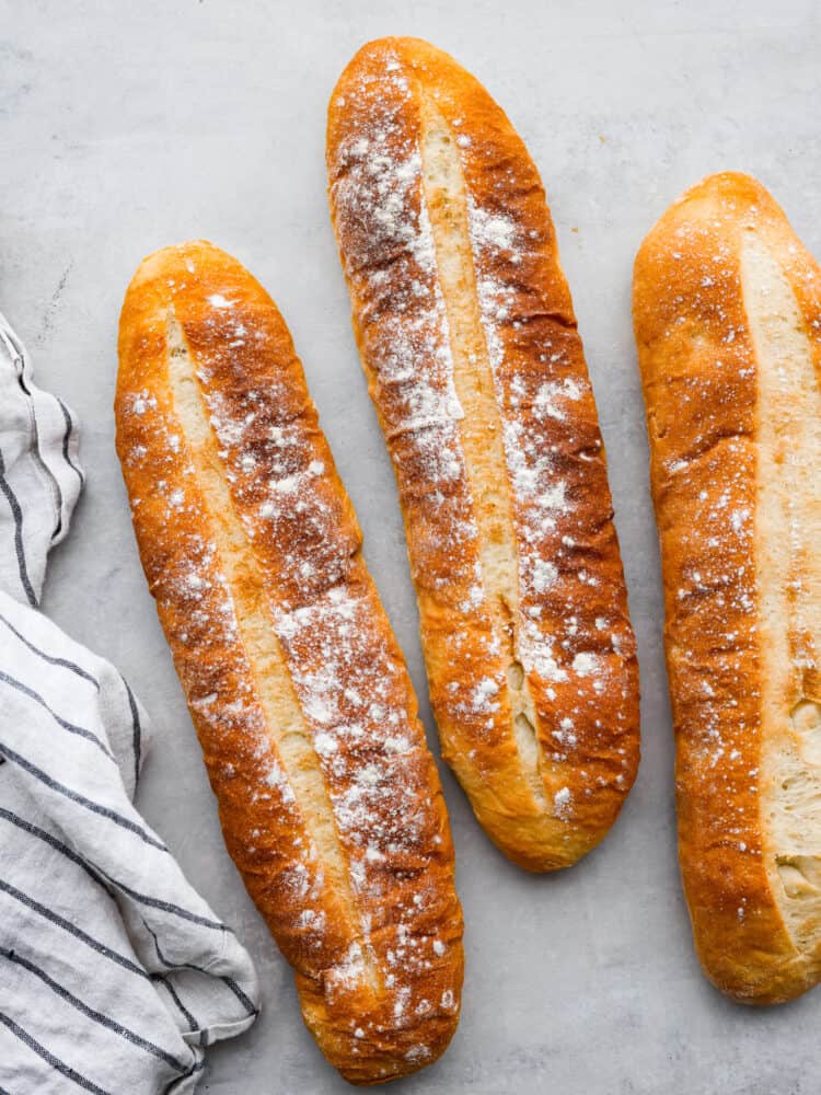 Cuban bread is a delightfully soft and fluffy bread that is shaped like a French baguette. It has a soft crust and the secret ingredient is lard which is what gives this bread great flavor and the perfect chew! Three loaves of cuban bread sitting on a counter top with a tea towel next to them.