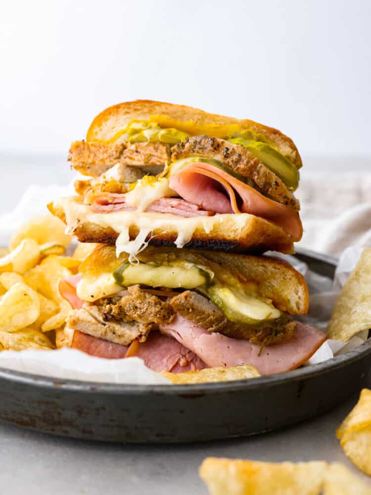 Cuban bread is a delightfully soft and fluffy bread that is shaped like a French baguette. It has a soft crust and the secret ingredient is lard which is what gives this bread great flavor and the perfect chew! Cuban bread cut into a sandwich with lots of meat and cheese and pickles in it, with chips off to the side.
