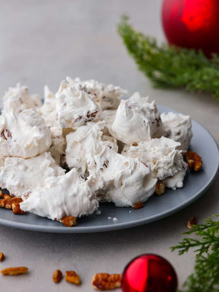 Divinity candy is an old fashioned Southern confection made from egg whites, corn syrup, and sugar. You will love the sweet soft texture of this classic holiday treat that is perfect for sharing with friends and neighbors. A pile of divinity on a gray plate. Pecans, greenery, and ornaments are styled around the plate.
