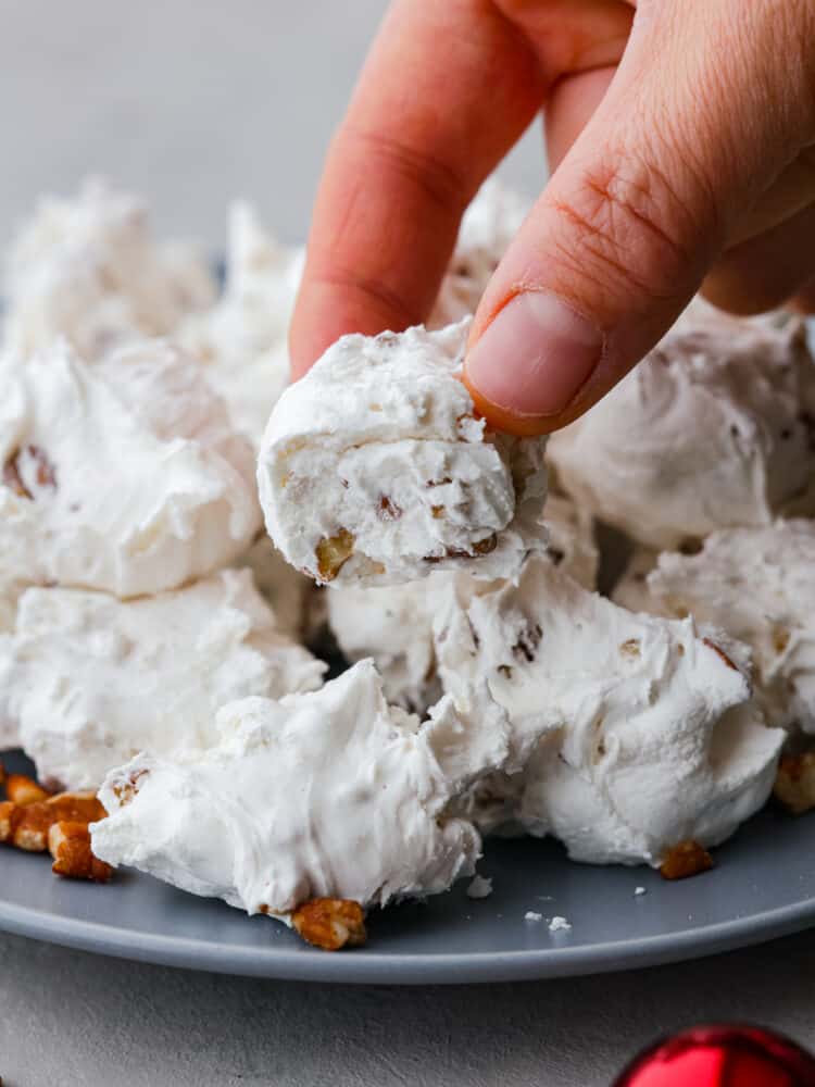 Divinity candy is an old fashioned Southern confection made from egg whites, corn syrup, and sugar. You will love the sweet soft texture of this classic holiday treat that is perfect for sharing with friends and neighbors. Close up view of a hand holding divinity with a bite out of it. Pecans are scattered around the plate of divinity.