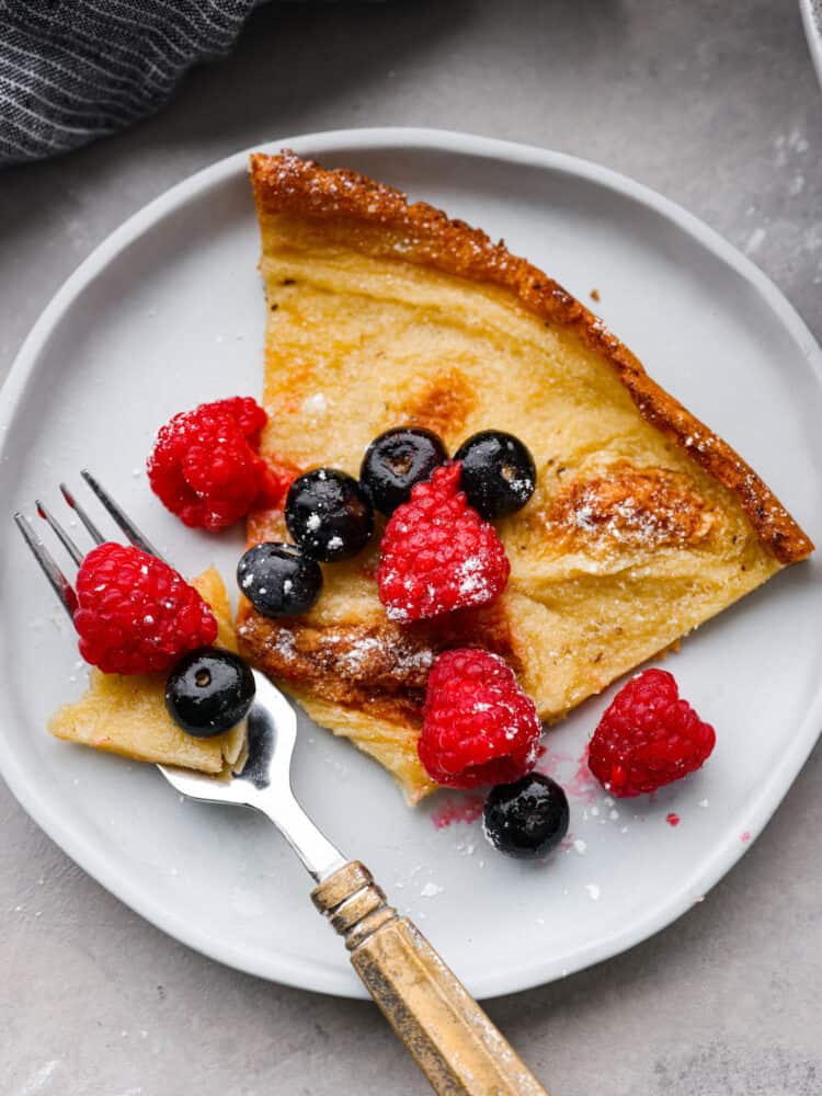 Light, fluffy, and deliciously oversized, this Dutch baby pancake is sure to please at the breakfast table! Add all of your favorite pancake toppings for a tasty treat the whole family will go crazy over. A slice of a Dutch baby on a white plate.