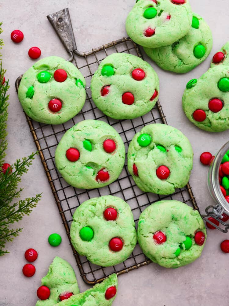 Grinch Cookies are a fun and festive twist on a classic chocolate chip cookie! Inspired by the popular Christmas movie, these soft and chewy cookies are colored Grinch-green and decorated with green and red M&Ms. Top view of Grinch cookies on a cooling rack. Extra cookies and M&Ms are scattered around the cookies on a gray counter top.