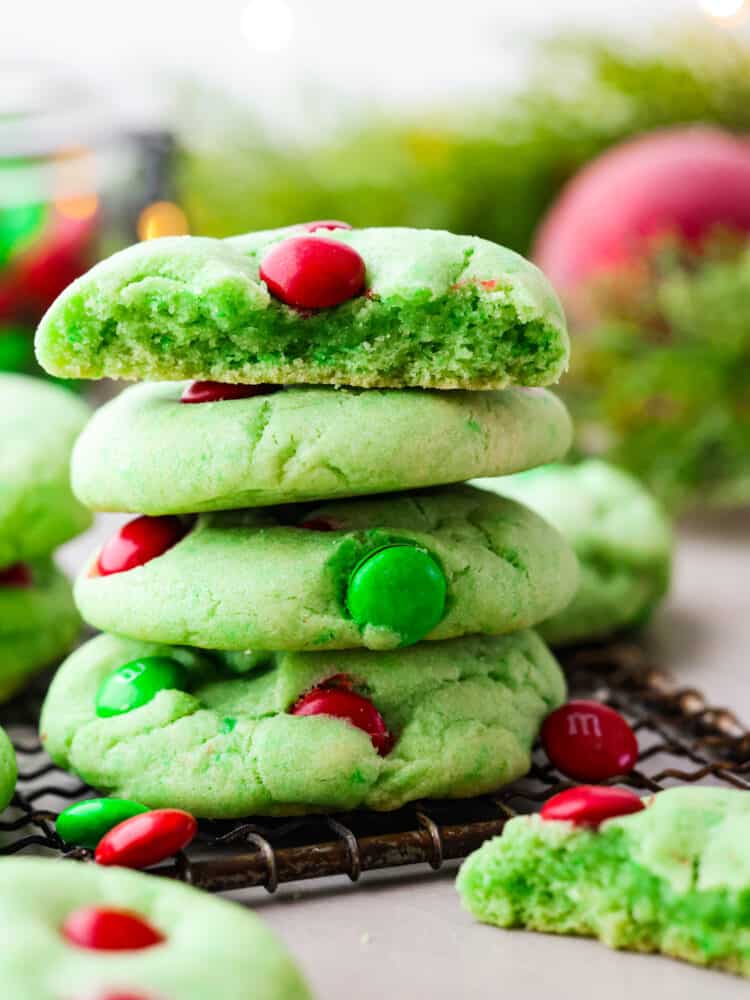 Grinch Cookies are a fun and festive twist on a classic chocolate chip cookie! Inspired by the popular Christmas movie, these soft and chewy cookies are colored Grinch-green and decorated with green and red M&Ms. Close up side view of stacked cookies on a cooling rack. A broken cookie and Christmas ornaments are styled next to the stack of Grinch cookies.