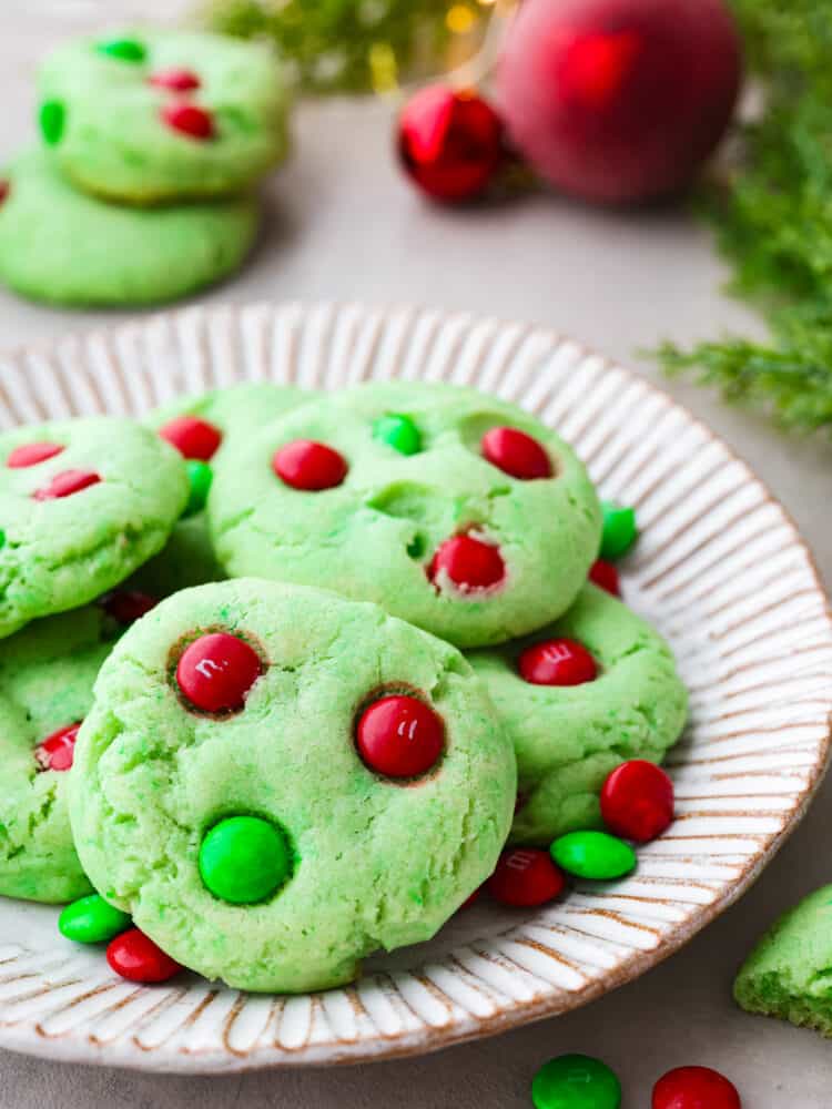 Grinch Cookies are a fun and festive twist on a classic chocolate chip cookie! Inspired by the popular Christmas movie, these soft and chewy cookies are colored Grinch-green and decorated with green and red M&Ms. Close up view of Grinch cookies stacked on a cream colored plate. Christmas ornaments, greenery and extra cookies are styled next to the plate.