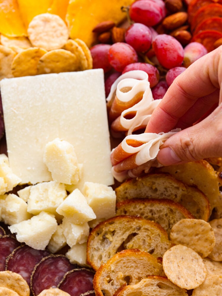 This classic Italian Charcuterie board is the perfect blend of savory and sweet. Loaded with nuts, dried fruit, cured meats, and a Caprese salad, this charcuterie board is perfect for your next get-together! A hand grabbing some meat from the charcuterie board.