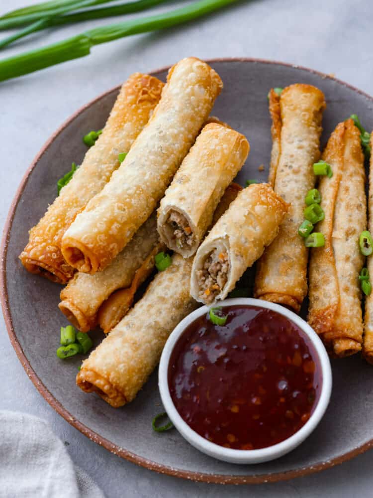 Lumpia is a delicious savory fried roll filled with ground pork, carrots, and cabbage. Traditionally found in the Philippines, lumpia is a perfect appetizer that is crispy and golden and served with dipping sauce on the side. Top view of lumpia on a gray plate with a side of dipping sauce. Chopped green onion on top for garnish.