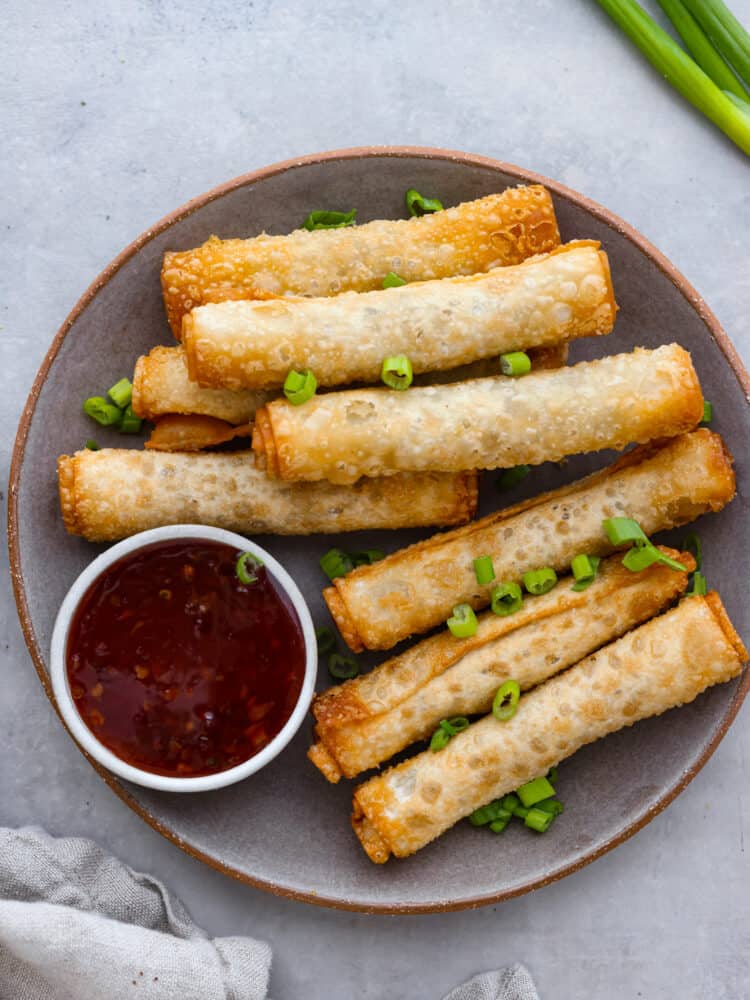 Lumpia is a delicious savory fried roll filled with ground pork, carrots, and cabbage. Traditionally found in the Philippines, lumpia is a perfect appetizer that is crispy and golden and served with dipping sauce on the side. Top view of lumpia on a gray plate with a side of dipping sauce. Chopped green onion on top for garnish.