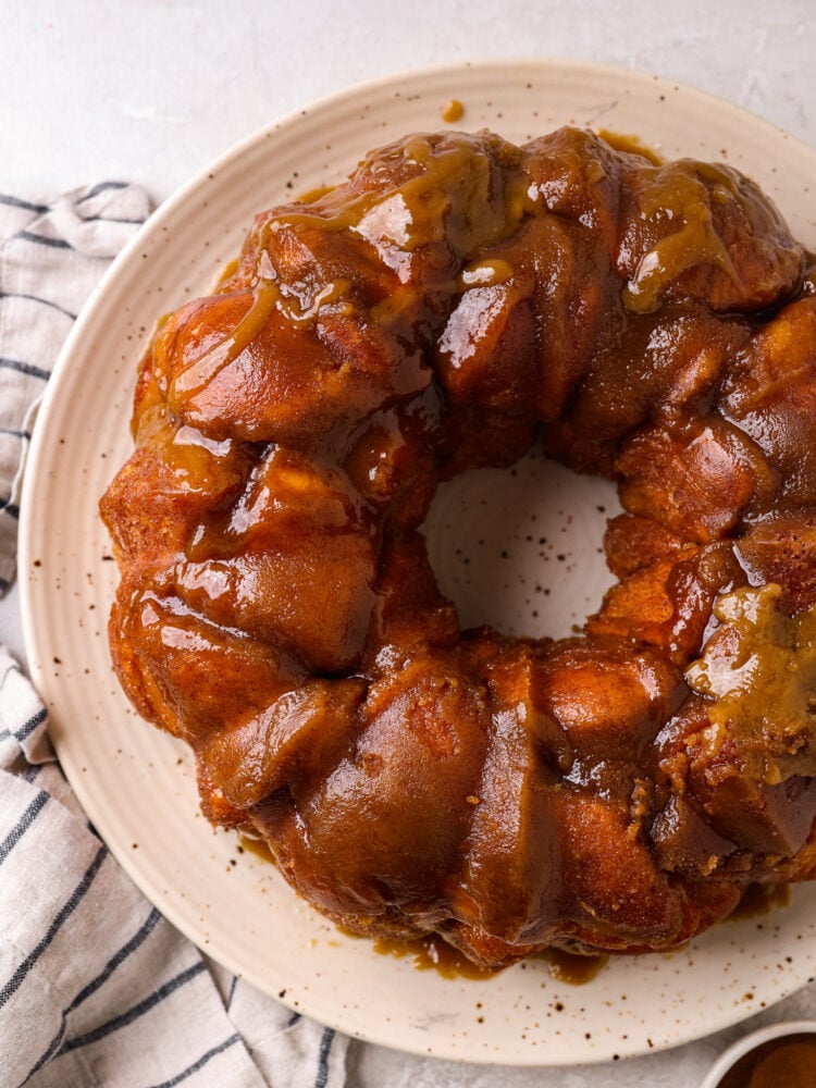 Soft and tender biscuits are covered in a gooey glaze with a sweet caramelized topping in this quick and easy monkey bread recipe! This decadent treat makes mornings or special events a little sweeter. Top-down view of monkey bread on a white plate.