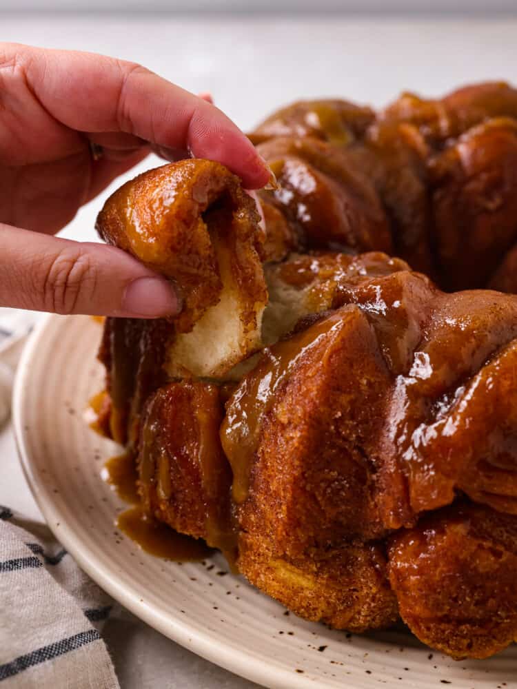 Soft and tender biscuits are covered in a gooey glaze with a sweet caramelized topping in this quick and easy monkey bread recipe! This decadent treat makes mornings or special events a little sweeter. A piece of monkey bread being pulled apart.