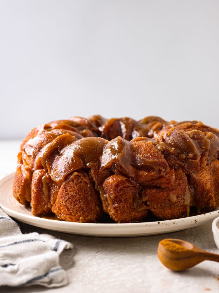 Soft and tender biscuits are covered in a gooey glaze with a sweet caramelized topping in this quick and easy monkey bread recipe! This decadent treat makes mornings or special events a little sweeter. Hero image of monkey bread on a white plate.