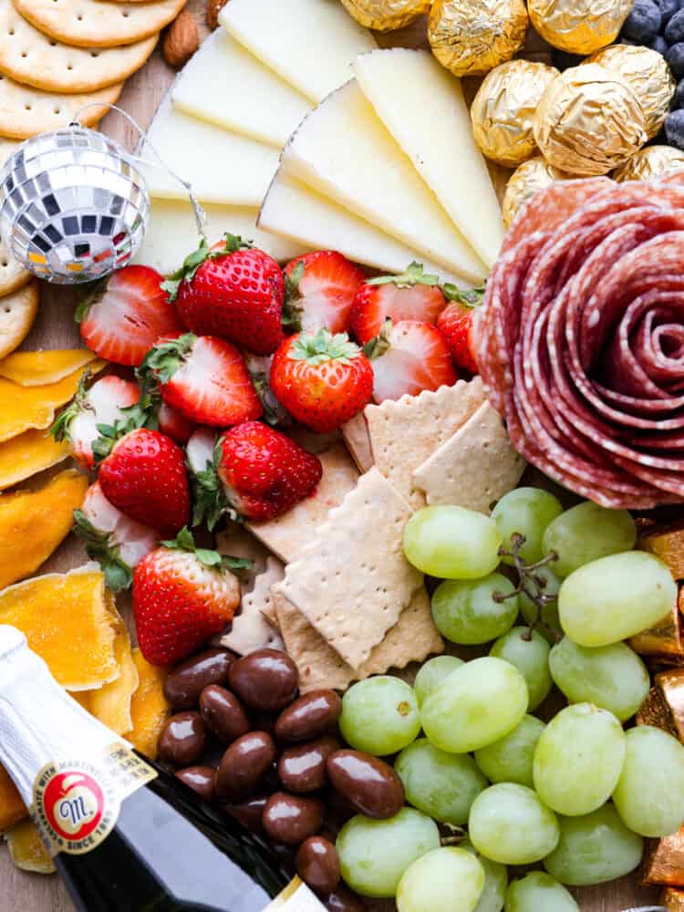 Throw a fun party and ring in the new year with this fantastic New Year's charcuterie board! Endless options of sweet and savory finger foods will keep your guests awake until midnight. Close up view of fruit, crackers, cheese, and salami rose on charcuterie board.