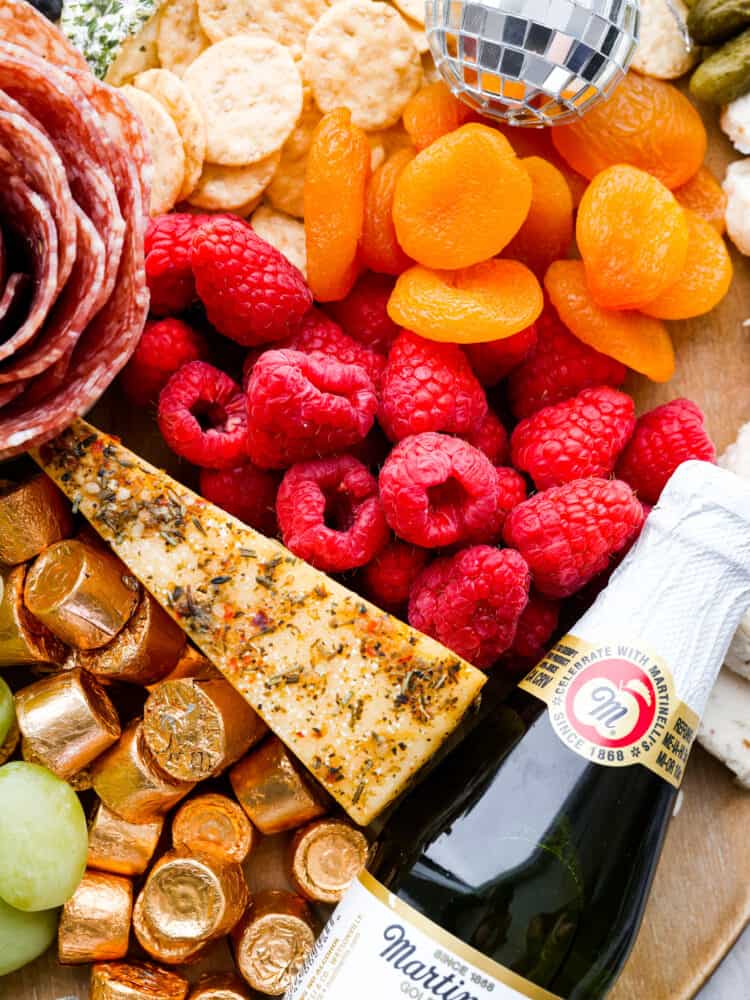 Throw a fun party and ring in the new year with this fantastic New Year's charcuterie board! Endless options of sweet and savory finger foods will keep your guests awake until midnight. Close up view of fruit, crackers, cheese, chocolate candy, and salami rose on charcuterie board. A miniature sparkling cider and disco ball is garnished on the board.