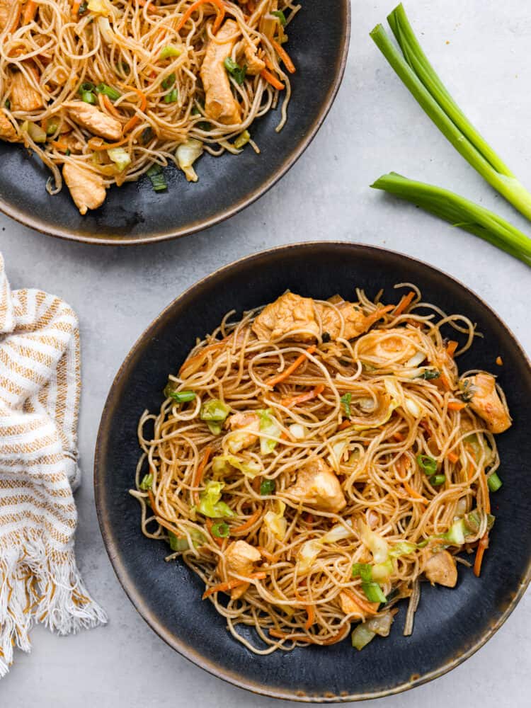 Pancit is a traditional Filipino stir-fry made with tender chicken, carrots, cabbage, and thin pancit noodles. The delicate rice noodles and savory sauce have made this meal a favorite for generations. Top view of pancit noodles into two black bowls. Green onion and a tan kitchen towel are next to the bowls.