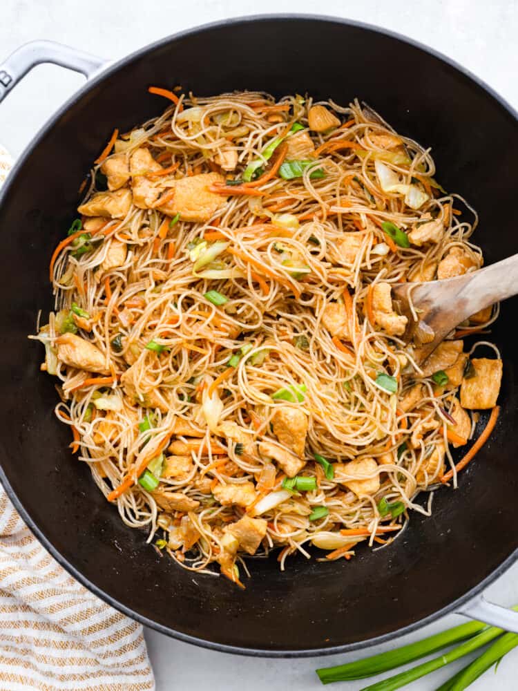 Pancit is a traditional Filipino stir-fry made with tender chicken, carrots, cabbage, and thin pancit noodles. The delicate rice noodles and savory sauce have made this meal a favorite for generations. Panict in a large black wok with a wooden serving spoon in the wok.