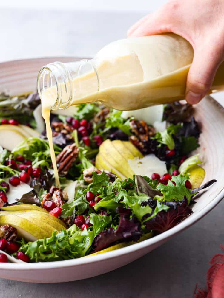 This pomegranate salad has just the right ingredients to go with any dish this holiday season! Packed with texture, different fruits, and delicious greens, you will be coming back for seconds (and probably even thirds)! A bottle of champagne vinaigrette being poured onto the salad.