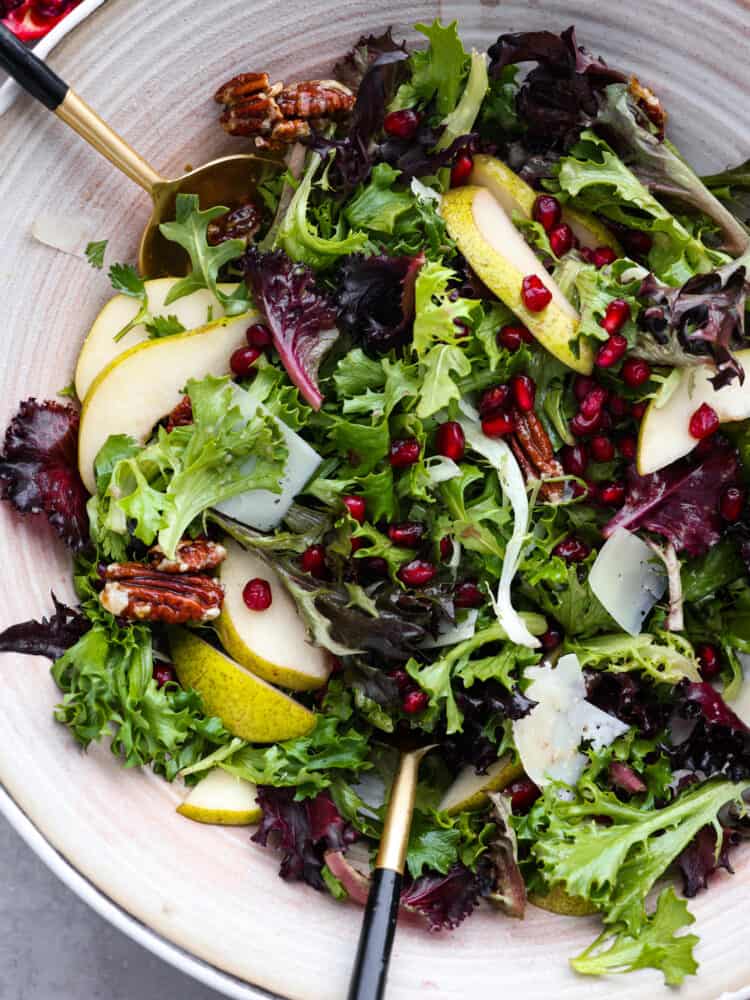 This pomegranate salad has just the right ingredients to go with any dish this holiday season! Packed with texture, different fruits, and delicious greens, you will be coming back for seconds (and probably even thirds)! A close up top view of a pomegranate salad with gold tongs.