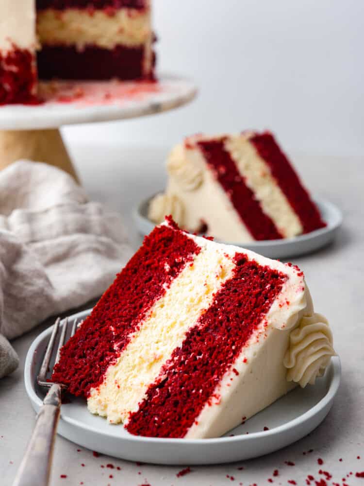 Can’t decide between red velvet cake and cheesecake? Why not both! This dreamy dessert is made with layers of moist red velvet cake and rich cheesecake topped with a luscious cream cheese frosting. A slice of cake (with another in the background) served on gray plates.