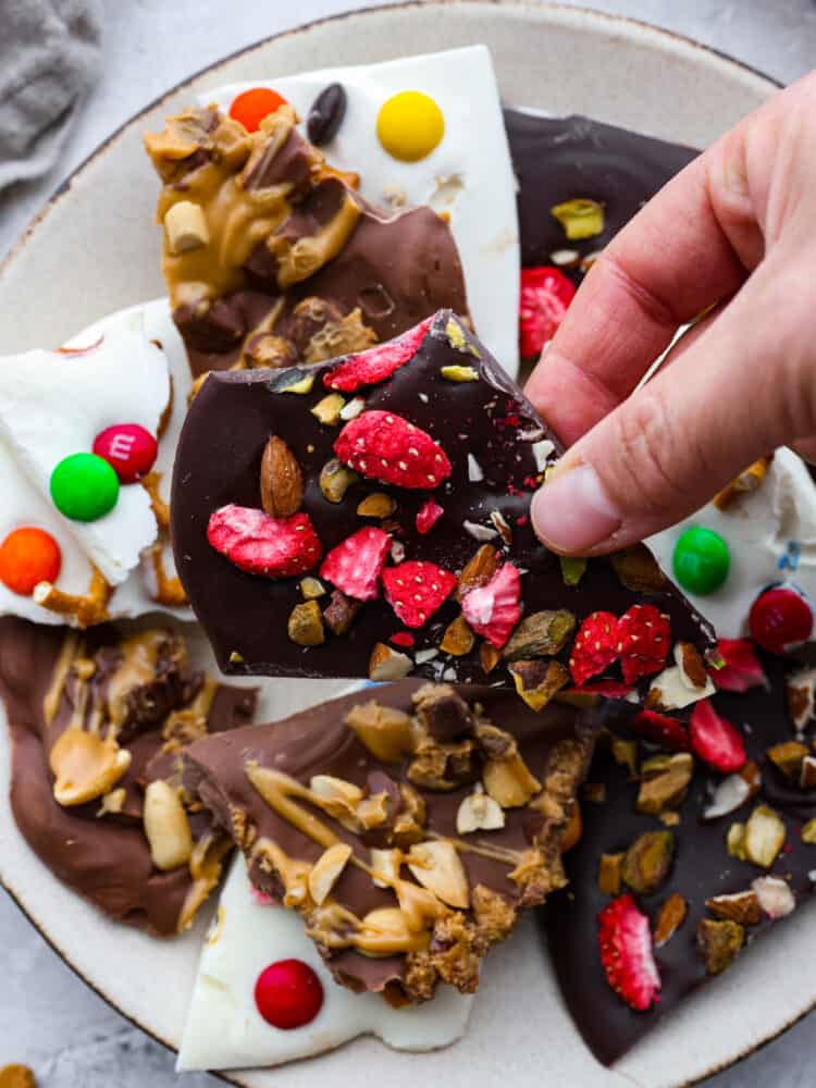 This delicious Chocolate Bark made withย Reynolds Wrapยฎ Heavy Duty Foilย will be the tastiest and simplest dessert you make all holiday season! It's quick and easy, and your entire family will go crazy over it! A plate of different kinds of chocolate bark with a hand grabbing a piece.