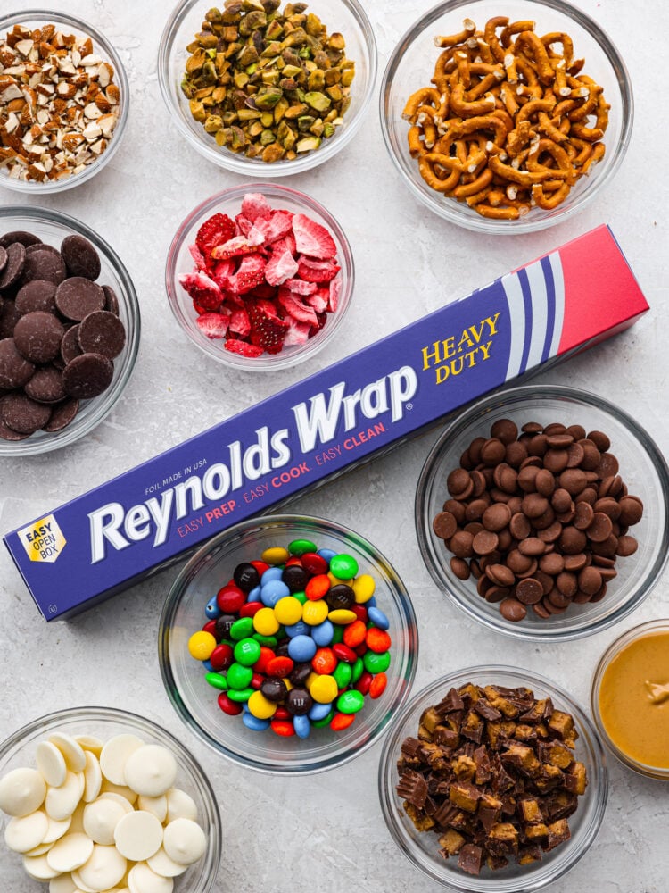This delicious Chocolate Bark made withย Reynolds Wrapยฎ Heavy Duty Foilย will be the tastiest and simplest dessert you make all holiday season! It's quick and easy, and your entire family will go crazy over it! Bowls of topping with chocolate, m &m's. pretzels, nuts and peanut butter with a Reynolds Wrap Foil package in the middle.