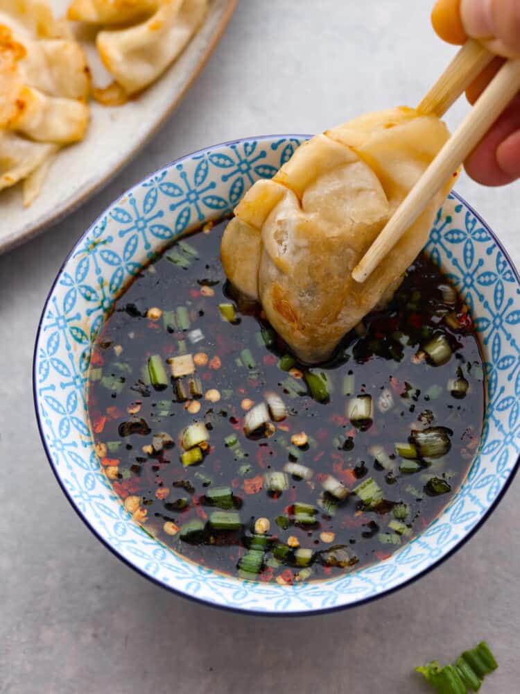 Potsticker sauce is easy to make at home and is the perfect dipping sauce for potstickers or any Asian appetizer! This delicious sauce is salty, tangy, and has a touch of sweetness to balance out the flavors. Close up view of potsticker sauce in a blue bowl. A potsticker is being dipped into the sauce with chopsticks.