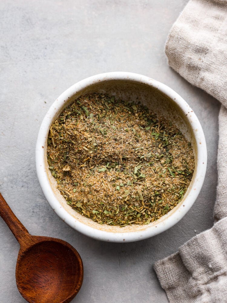 Look no further for the BEST turkey seasoning recipe you will ever make! The combination of herbs and spices in this rub will flavor your turkey to perfection and it will be your go-to turkey rub. Top view of turkey seasoning in a small white bowl with a wooden spoon and tan kitchen towel next to the bowl.